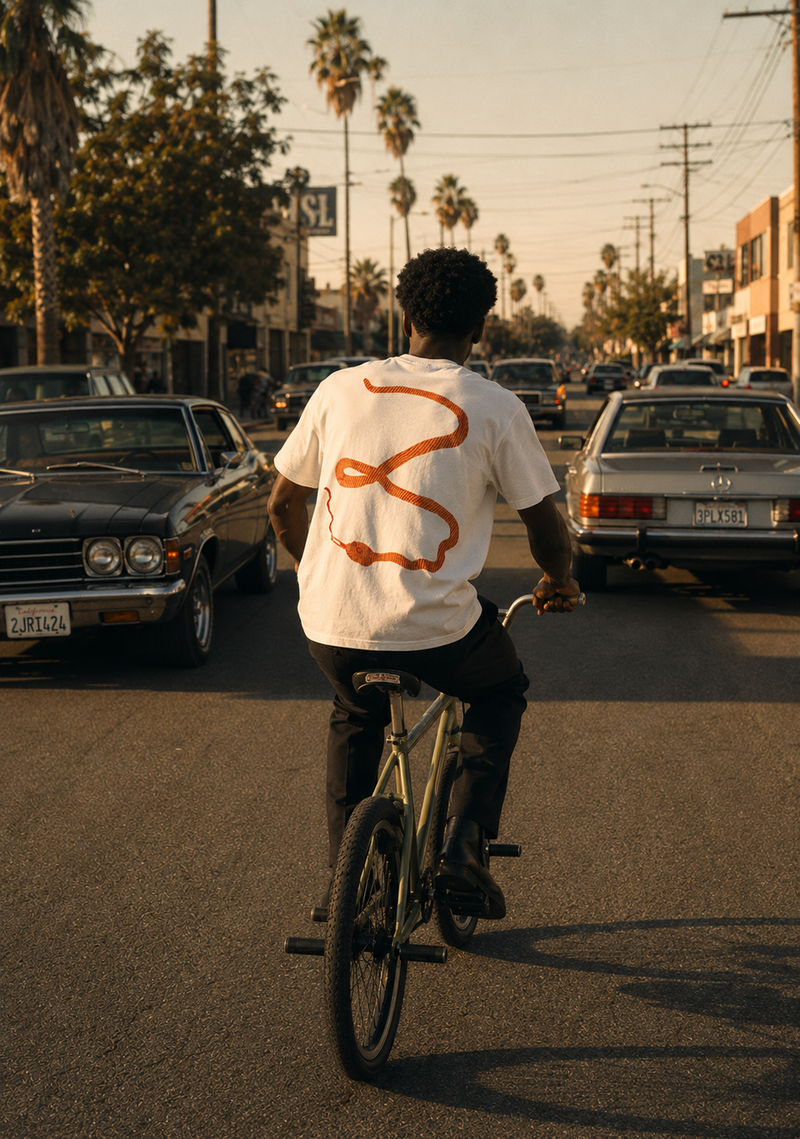 Back of BISHOP white tee - orange snake graphic on model riding BMX through LA traffic at golden hour, EUPHORIC collection by Nostalgic Misfits