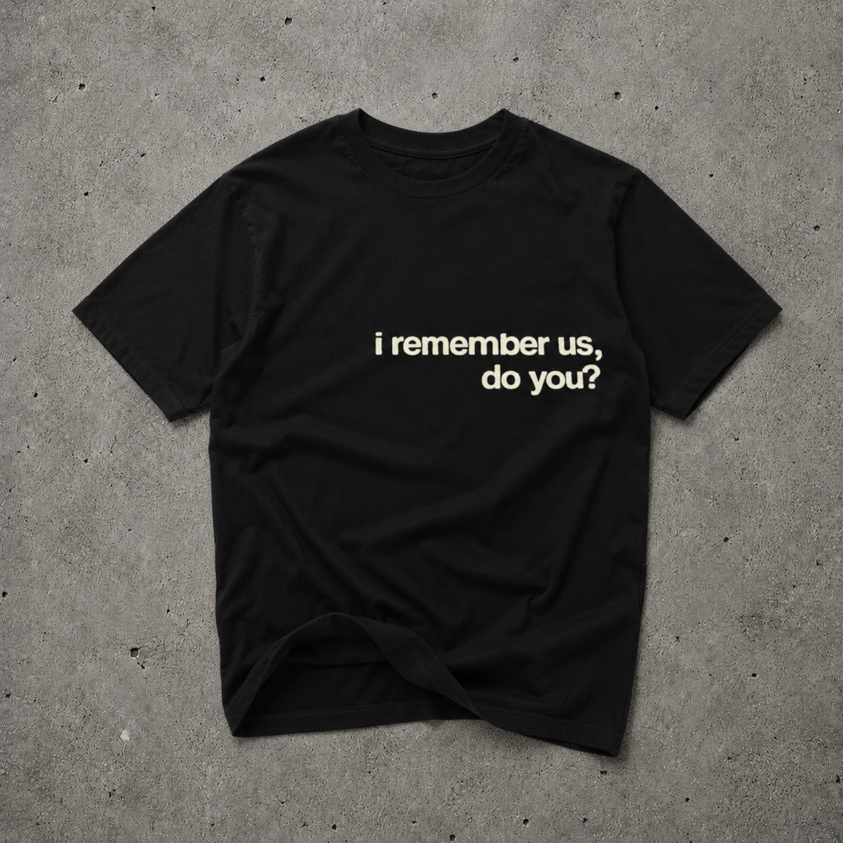 Black unisex t-shirt with cream bold text print reading "i remember us, do you?", flat lay on concrete background