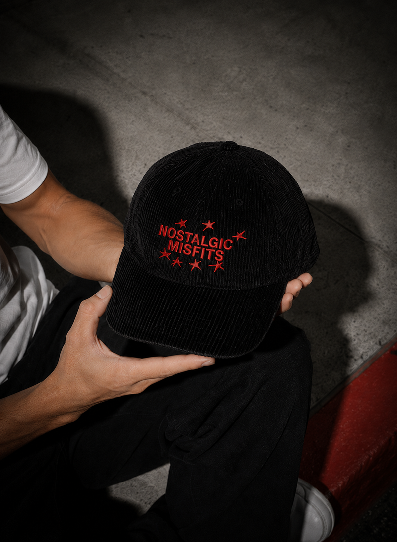 Black corduroy Nostalgic Misfits cap held in hands against a concrete wall — top-down view showing red embroidered NOSTALGIC MISFITS logo with stars on the front panel