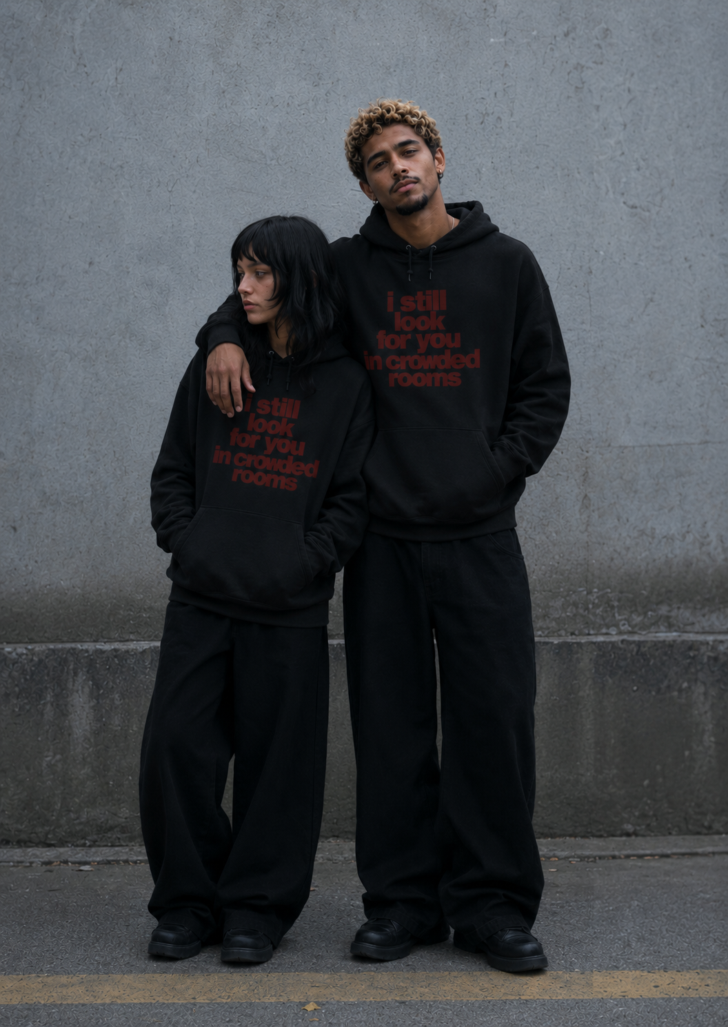 A woman and a man standing close together against a grey concrete wall, both wearing matching black Nostalgic Misfits hoodies with dark red "i still look for you in crowded rooms" print