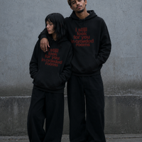 A woman and a man standing close together against a grey concrete wall, both wearing matching black Nostalgic Misfits hoodies with dark red "i still look for you in crowded rooms" print