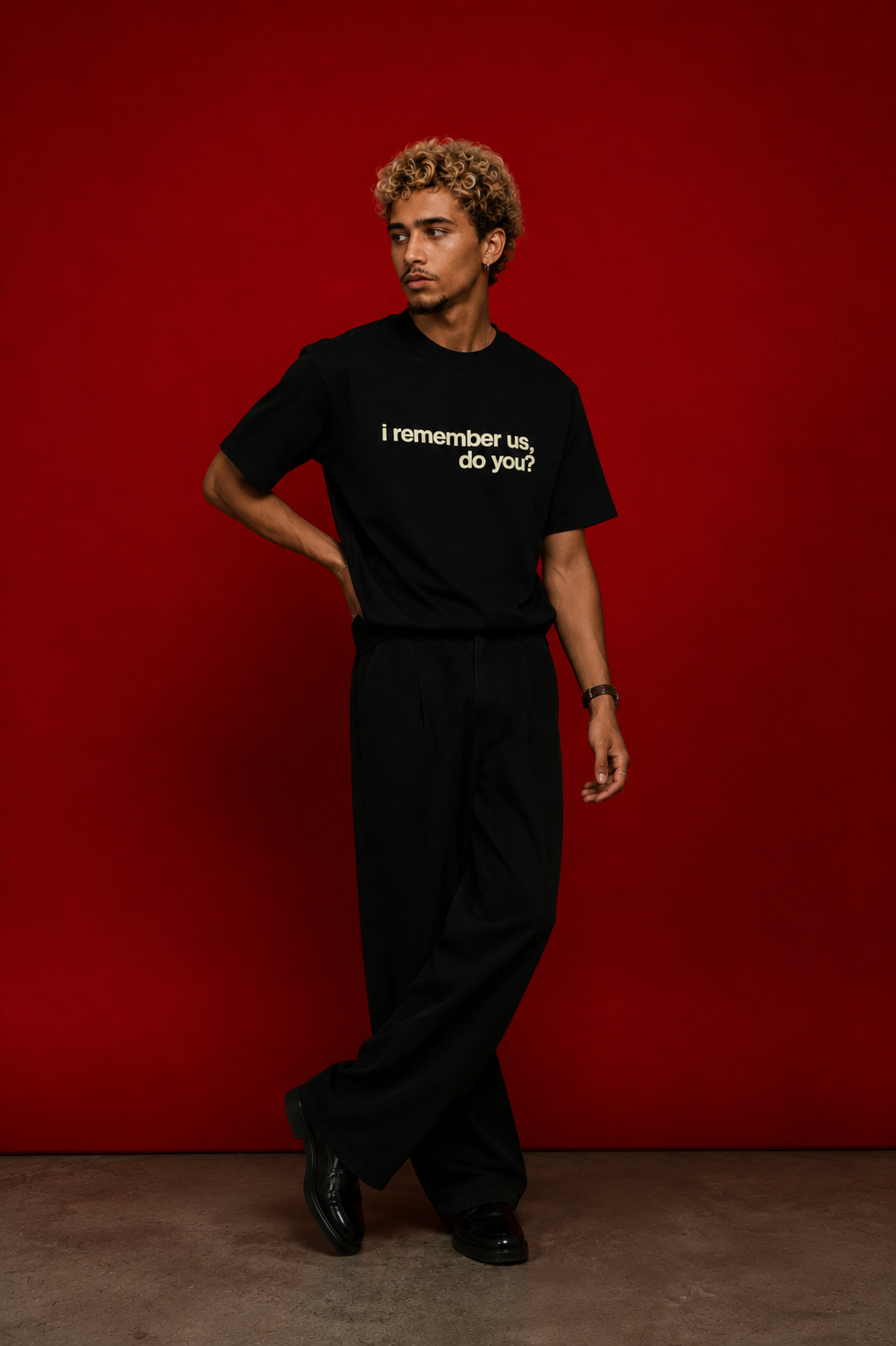 Male model wearing black "i remember us, do you?" t-shirt standing against deep red background, styled with wide-leg black trousers and black boots