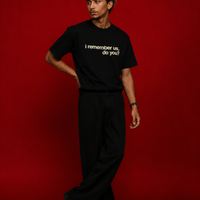 Male model wearing black "i remember us, do you?" t-shirt standing against deep red background, styled with wide-leg black trousers and black boots