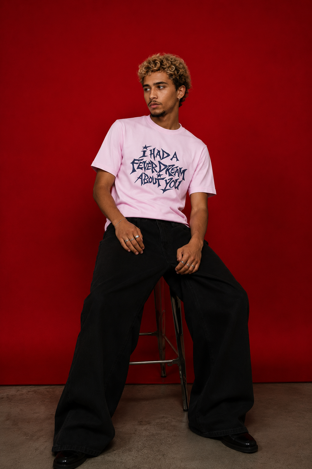 Male model wearing pink "I Had a Fever Dream About You" t-shirt seated against red background, styled with wide-leg black trousers
