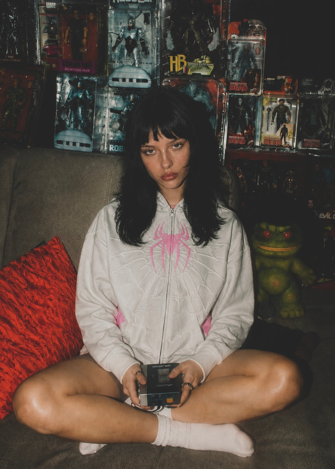 Nostalgic Misfits Ghost Zip white pink spidey hoodie female model sitting on sofa surrounded by vintage action figures collector lifestyle editorial shot