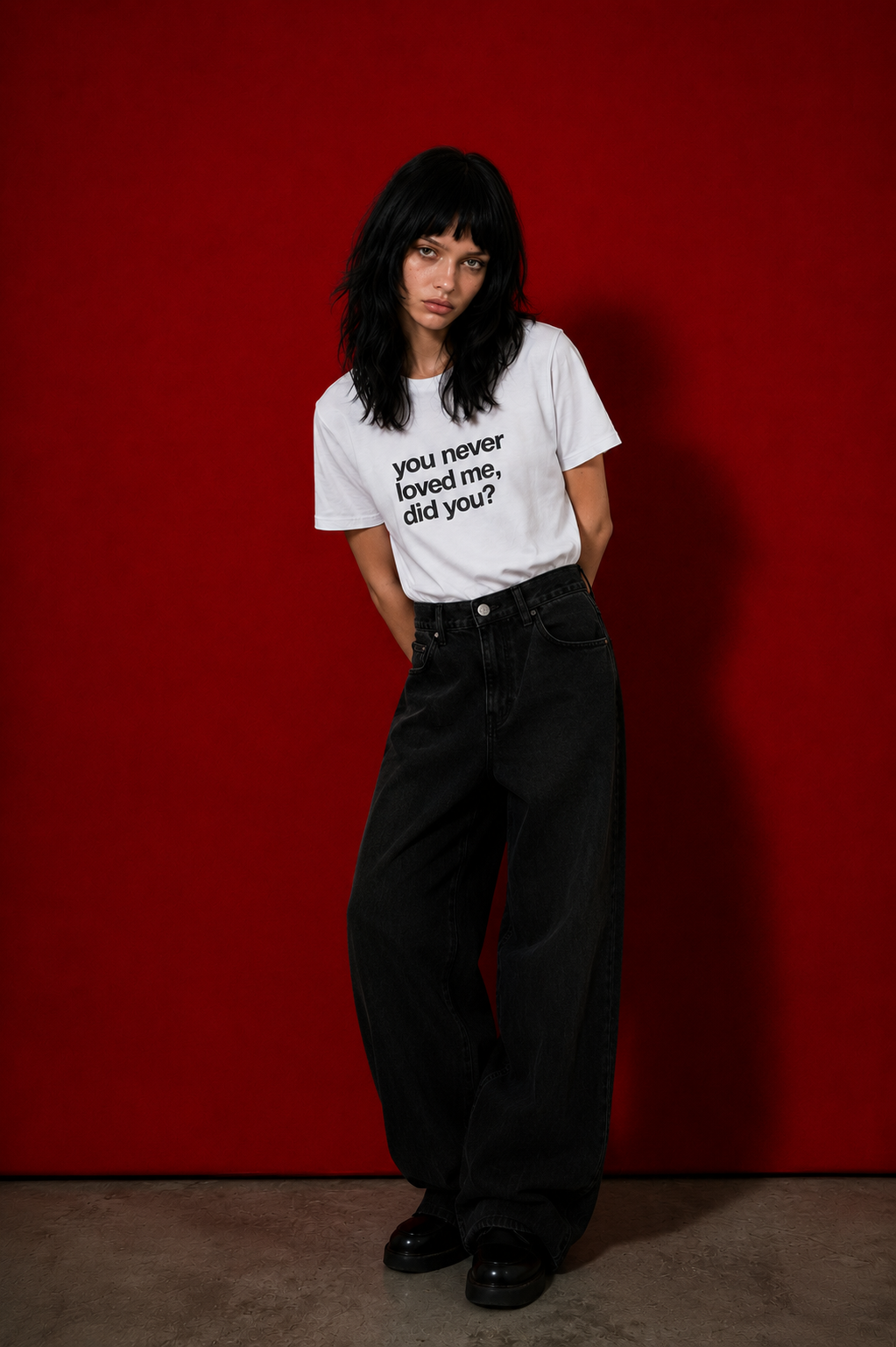 Female model wearing white "you never loved me, did you?" t-shirt standing against deep red background, styled with wide-leg black denim jeans and black boots