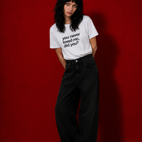 Female model wearing white "you never loved me, did you?" t-shirt standing against deep red background, styled with wide-leg black denim jeans and black boots
