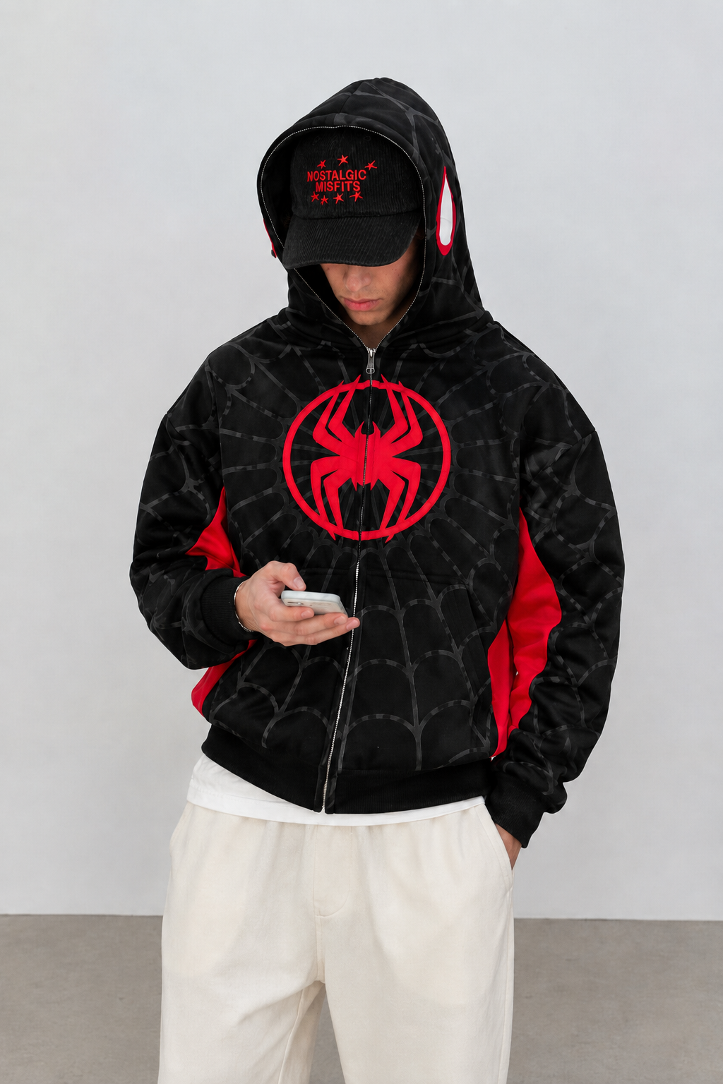 Nostalgic Misfits Miles Zip black red spidey hoodie model wearing Nostalgic Misfits black cap cream sweatpants holding phone hood up streetwear editorial look
