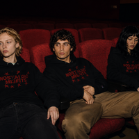 Three young people wearing matching black Nostalgic Misfits x Champion hoodies, sitting side by side in red cinema seats in a darkened movie theatre, looking forward with a quiet, melancholic expression