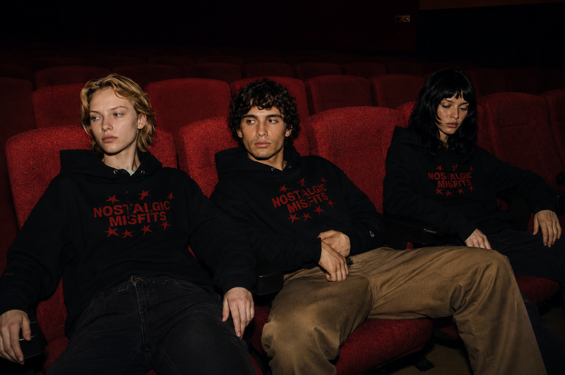 Three young people wearing matching black Nostalgic Misfits x Champion hoodies, sitting side by side in red cinema seats in a darkened movie theatre, looking forward with a quiet, melancholic expression