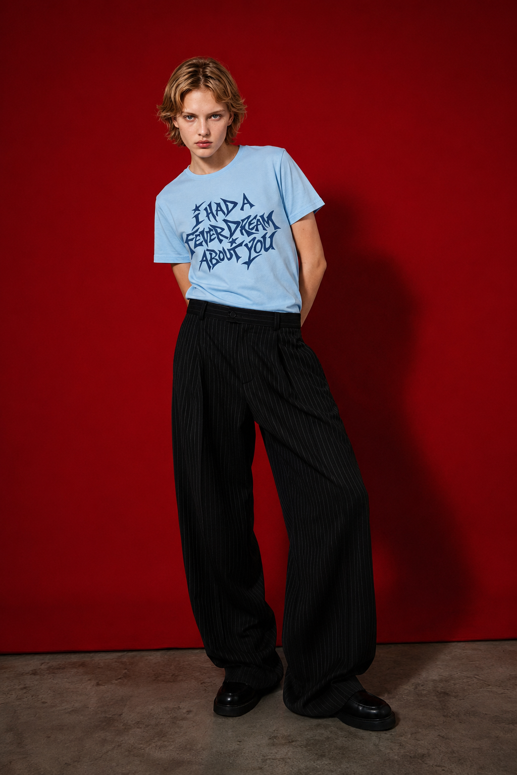 Female model wearing baby blue "I Had a Fever Dream About You" t-shirt standing against red background, styled with wide-leg pinstripe black trousers