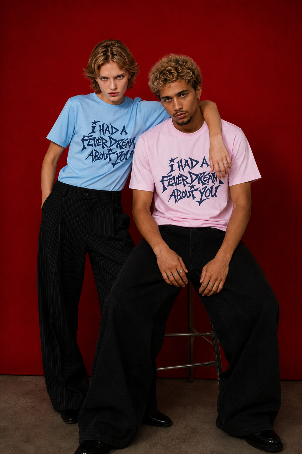 Two models wearing "I Had a Fever Dream About You" t-shirts against a deep red background — one in baby blue, one in pink, with navy graphic print
