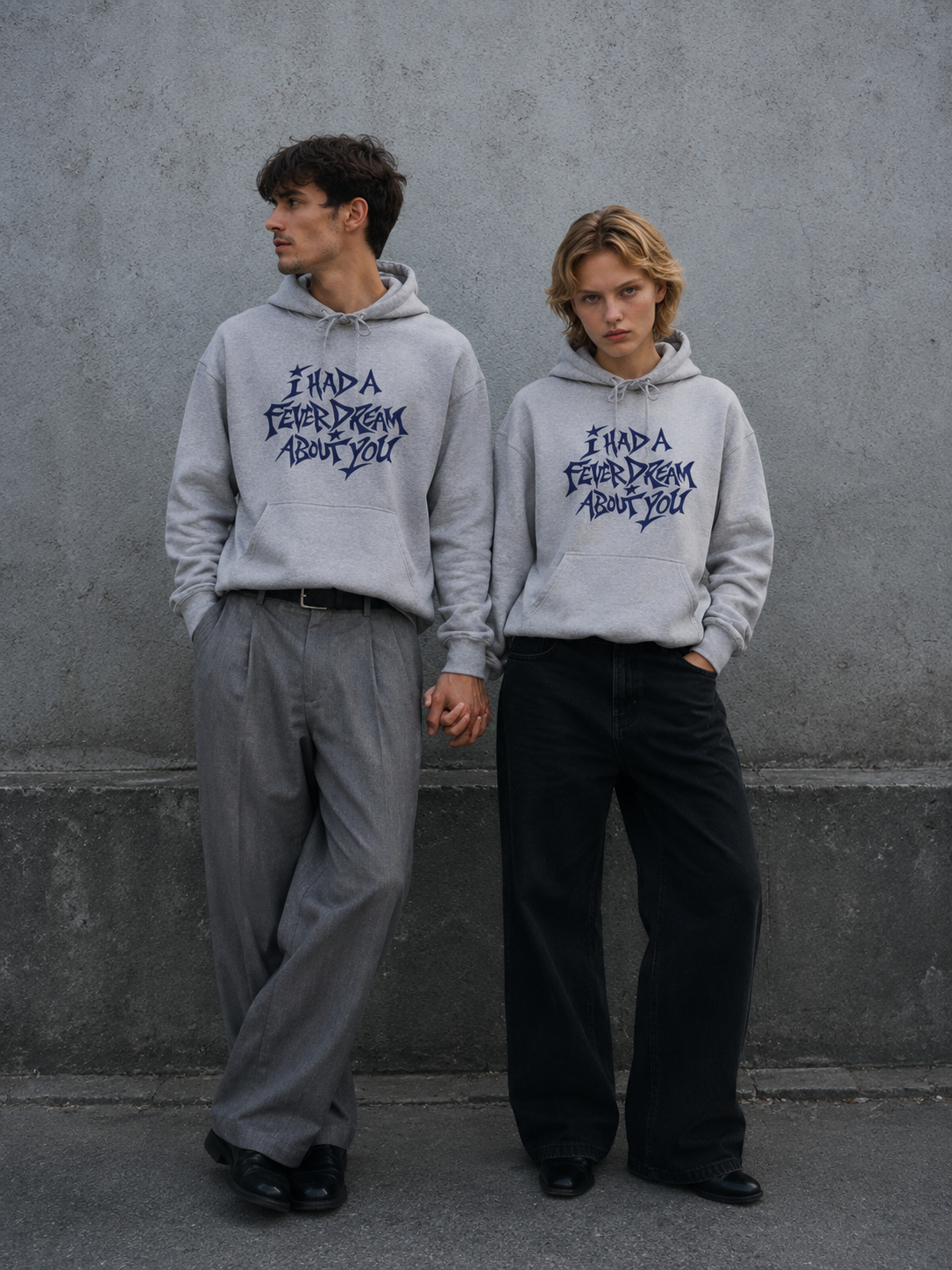 A man and a woman standing side by side against a grey concrete wall, both wearing matching grey Nostalgic Misfits hoodies with navy "I Had A Fever Dream About You" print, holding hands