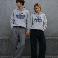 A man and a woman standing side by side against a grey concrete wall, both wearing matching grey Nostalgic Misfits hoodies with navy "I Had A Fever Dream About You" print, holding hands