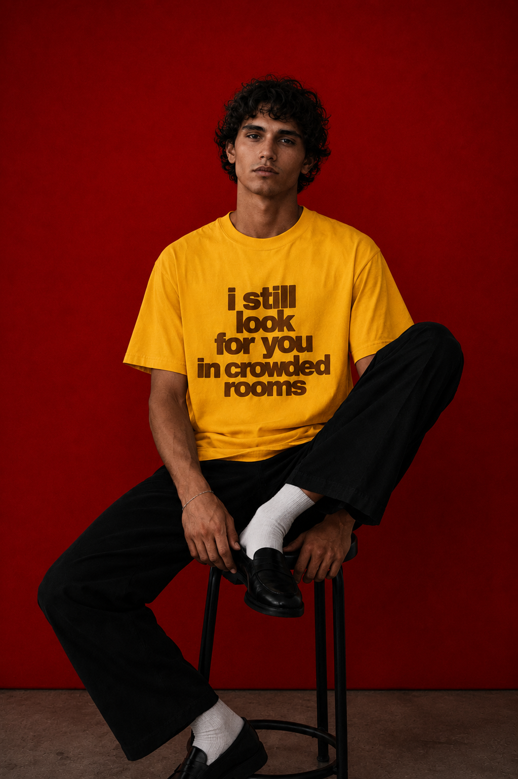Male model wearing yellow "i still look for you in crowded rooms" t-shirt seated on a stool against deep red background, styled with black wide-leg trousers and white socks with loafers