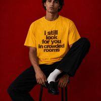 Male model wearing yellow "i still look for you in crowded rooms" t-shirt seated on a stool against deep red background, styled with black wide-leg trousers and white socks with loafers
