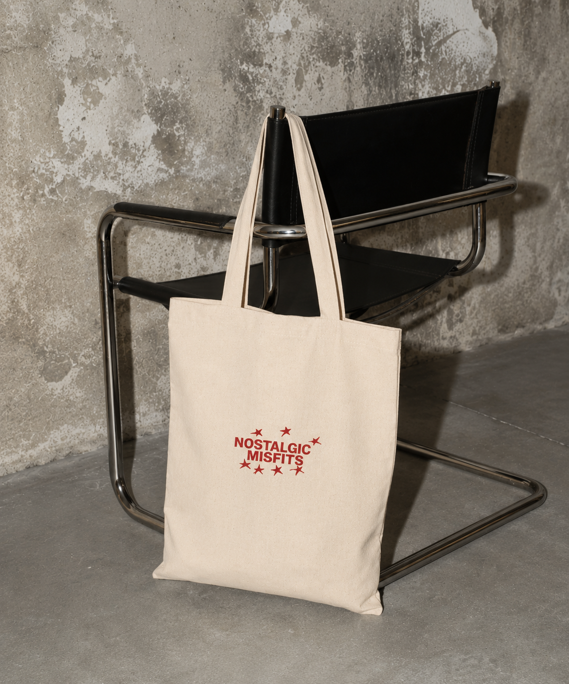 atural canvas Nostalgic Misfits tote bag hanging on a black leather chair against a weathered concrete wall — back view showing dark brown bold stacked text "i still look for you in crowded rooms"