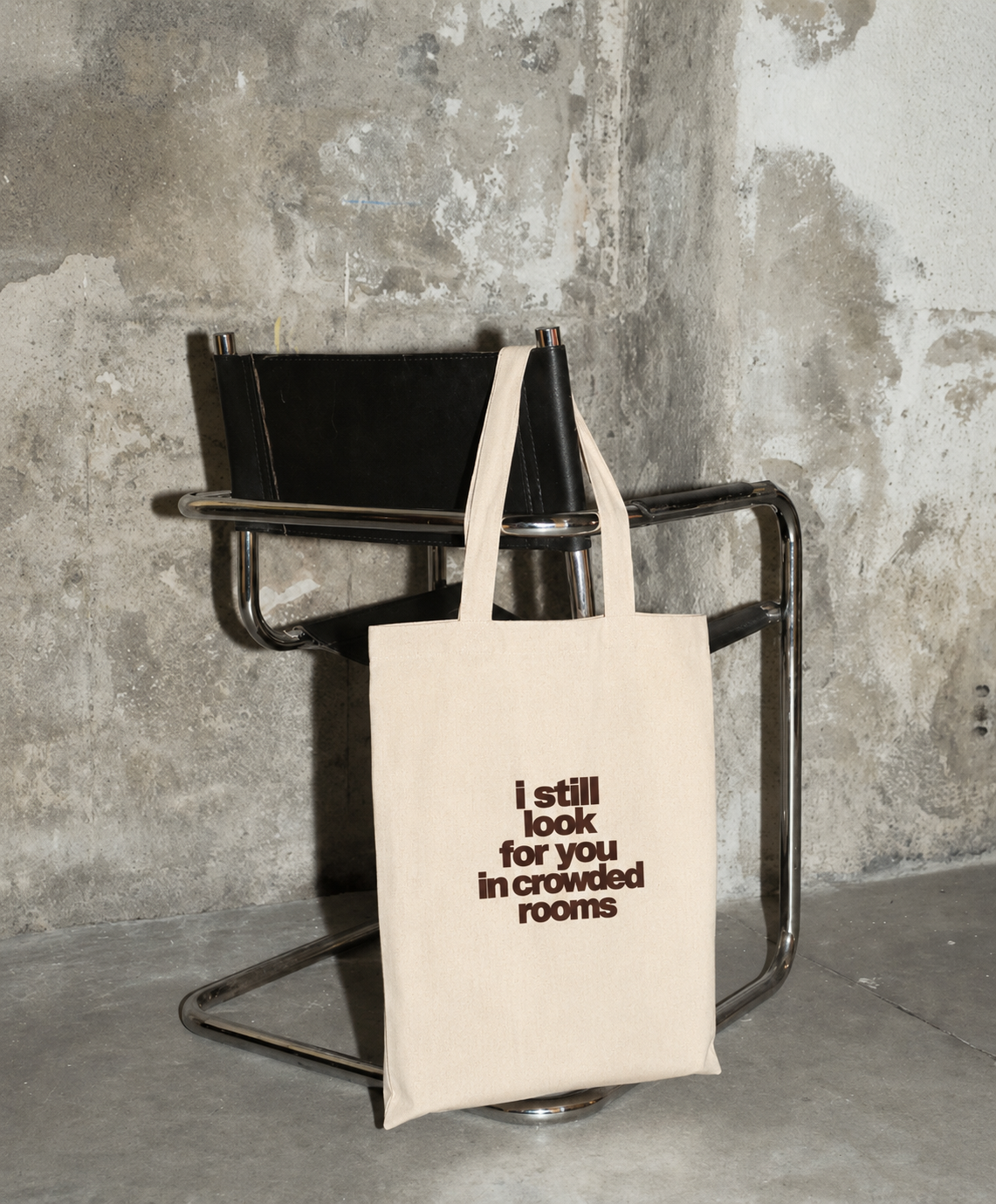 Natural canvas Nostalgic Misfits tote bag leaning against a black leather chair on concrete floor — front view showing red NOSTALGIC MISFITS star logo print
