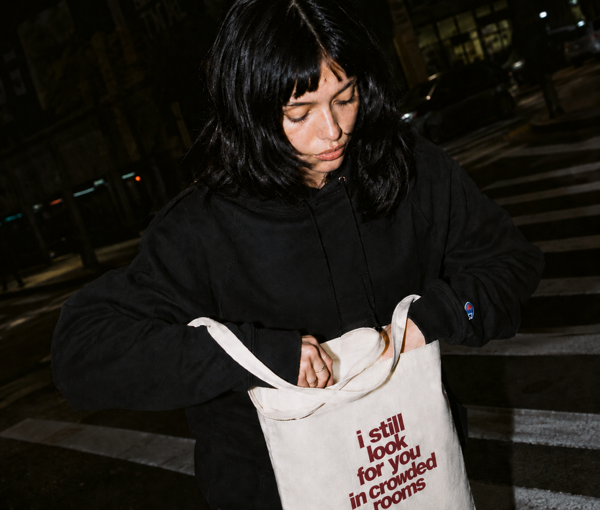Young woman with dark hair looking down into a natural canvas tote bag on a city street at night, wearing a black Nostalgic Misfits x Champion hoodie — bag shows "i still look for you in crowded rooms" print in dark red