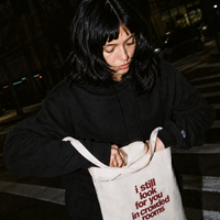Young woman with dark hair looking down into a natural canvas tote bag on a city street at night, wearing a black Nostalgic Misfits x Champion hoodie — bag shows "i still look for you in crowded rooms" print in dark red