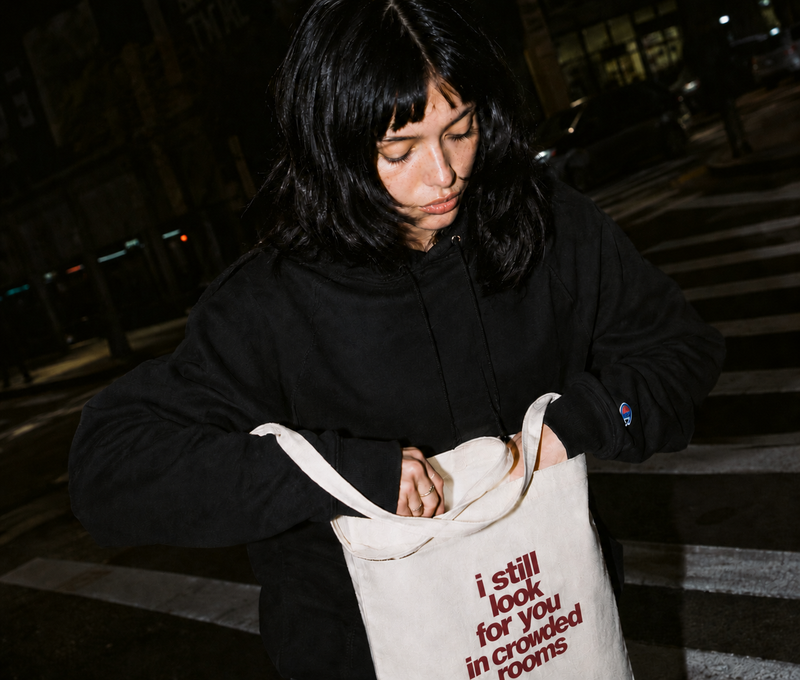Young woman with dark hair looking down into a natural canvas tote bag on a city street at night, wearing a black Nostalgic Misfits x Champion hoodie — bag shows "i still look for you in crowded rooms" print in dark red