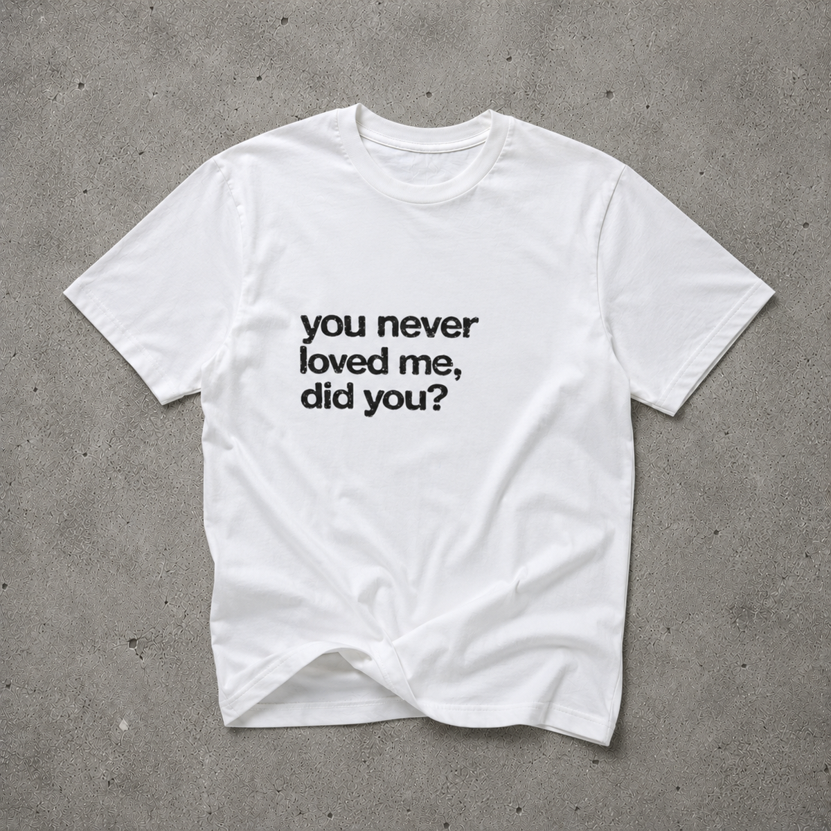 White unisex t-shirt with black bold text print reading "you never loved me, did you?", flat lay on concrete background