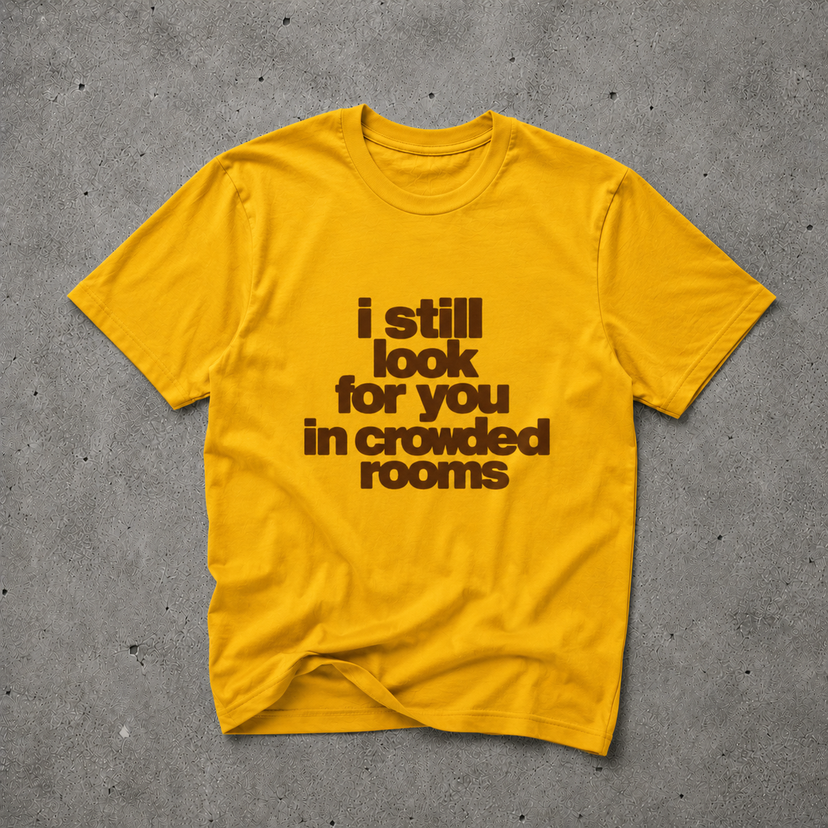 Yellow unisex t-shirt with brown bold text print reading "i still look for you in crowded rooms", flat lay on concrete background