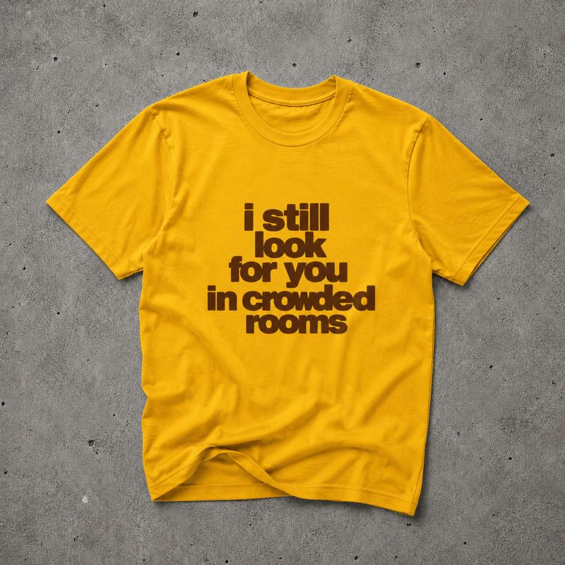 Yellow unisex t-shirt with brown bold text print reading "i still look for you in crowded rooms", flat lay on concrete background